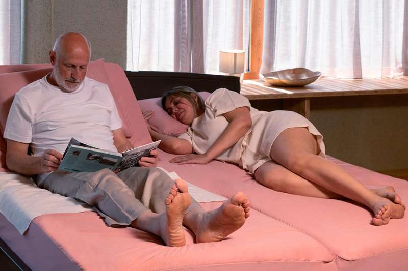 Hombre mayor leyendo en la cama con empapador visible y mujer descansando