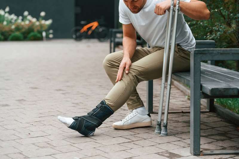 Hombre con bota ortopédica y muletas sentado en banco durante la recuperación de una fractura de tobillo